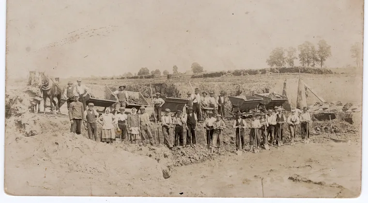 Sepia group photograph of around thirty labourers with shovels and wheelbarrows at an excavation site, with a horse cart and a flag