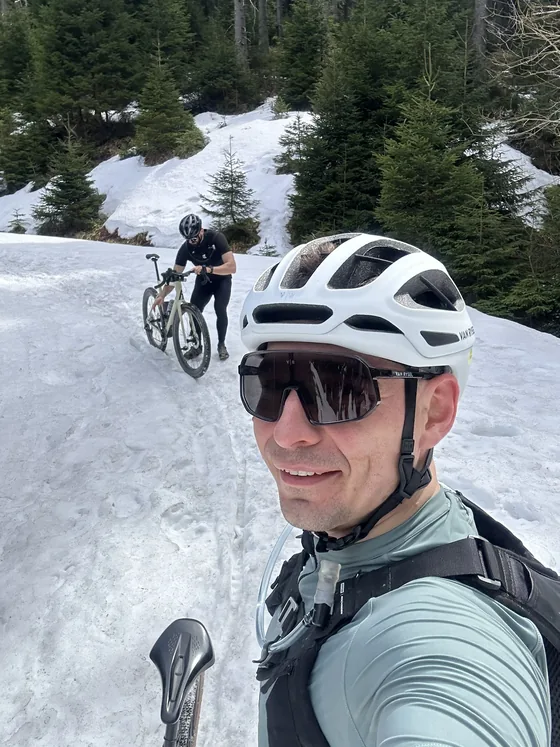 A snow-covered forest road with two gravel bikes, one rider pushing uphill in the background