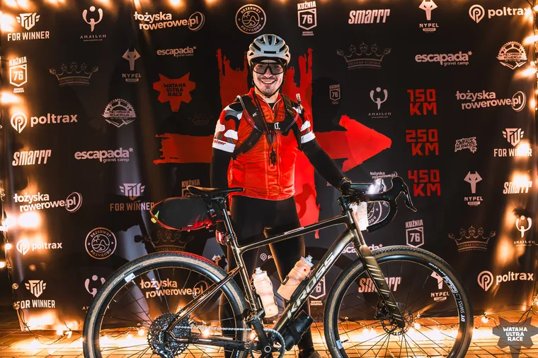 Marcin Wadas in race kit standing with his loaded gravel bike in front of the Wataha Ultra Race backdrop