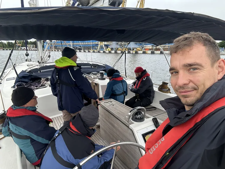 Six crew in foul-weather gear in the cockpit of a yacht under the bimini, one of them at the wheel, harbour cranes in the background