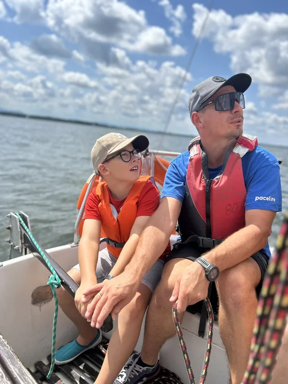 Marcin Wadas and a child, both in buoyancy aids, sitting on the side deck of a yacht on a sunny day