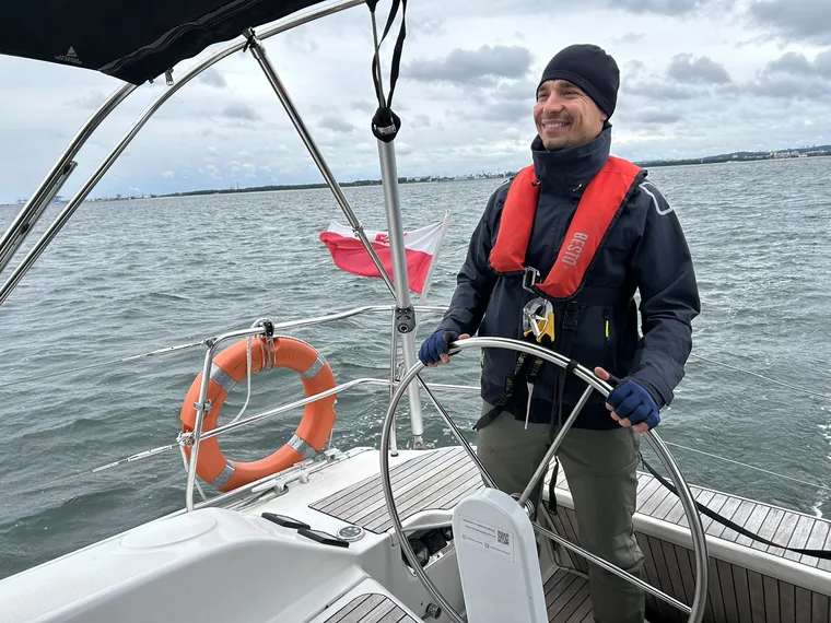 Marcin Wadas standing at the wheel of a yacht in offshore kit and an inflatable lifejacket, the Polish ensign flying and open grey water behind him