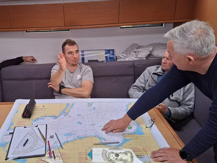 Crew gathered around the saloon table of a yacht while the skipper points at a paper chart spread out with dividers, a plotter and a pencil