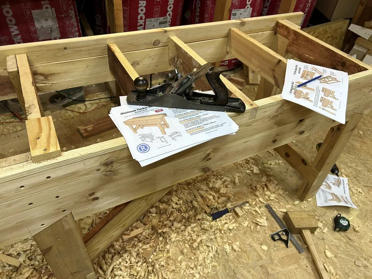 A partly built timber workbench frame with printed plans and a hand plane resting on top, wood shavings on the floor