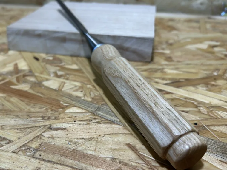 A chisel with a hand-shaped ash handle resting on a workbench beside a pale offcut